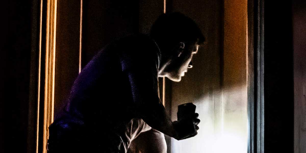 A person leans towards a door, holding a flashlight, with shadows and warm light illuminating the scene.