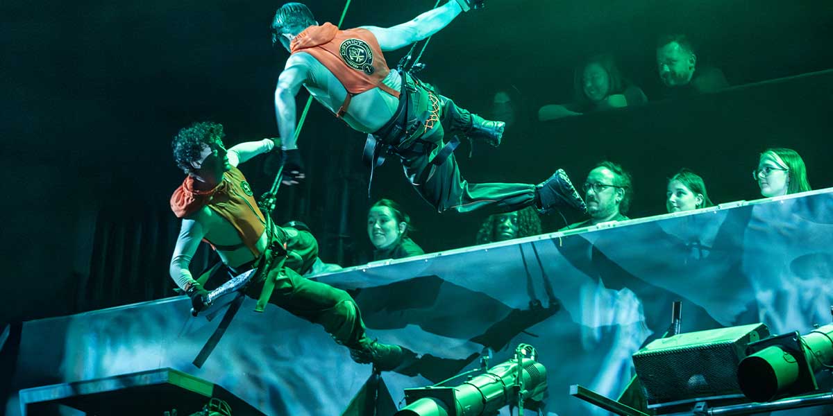 A dramatic scene from "The Hunger Games: On Stage" featuring characters in elaborate costumes against a dark backdrop.