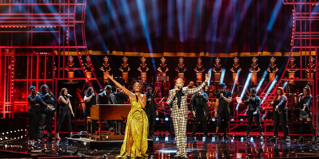 Beverley Knight and Billy Porter hosting the Olivier Awards 2025. They are standing on stage at the Royal Albert Hall. Beverley wears a yellow print dress and Billy wears a brown patterned suit.