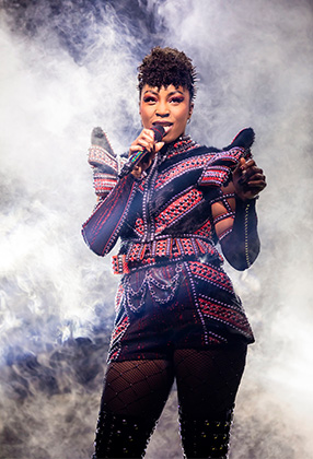 A performer in a striking black and red costume sings confidently amidst a smoky backdrop.