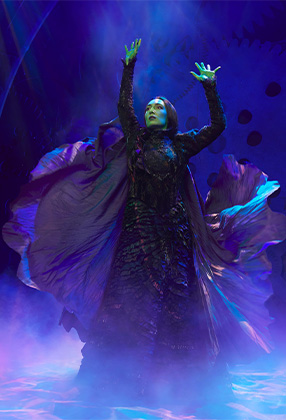 A performer in a flowing black costume raises their arms dramatically against a misty, colourful background.