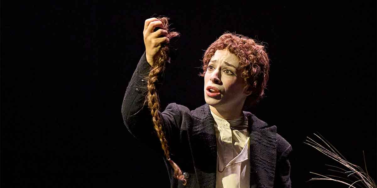 Amy Hack as Yentl holds a long braid, expressing emotion against a dark background.