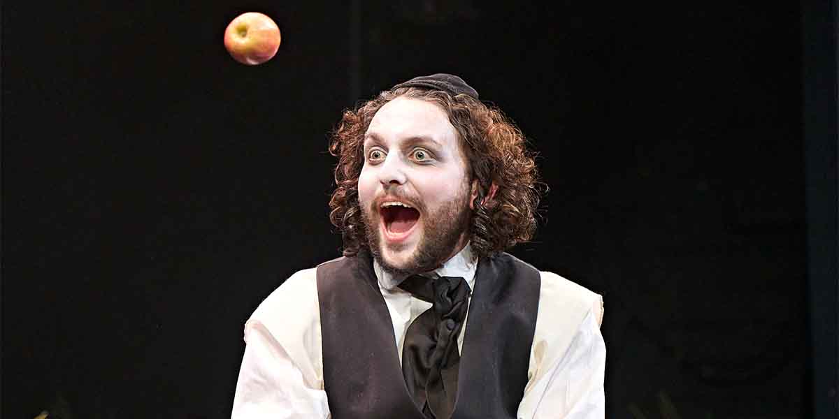 Ashley Margolis as Avigdor, with curly hair and white face paint, excitedly catches an apple.