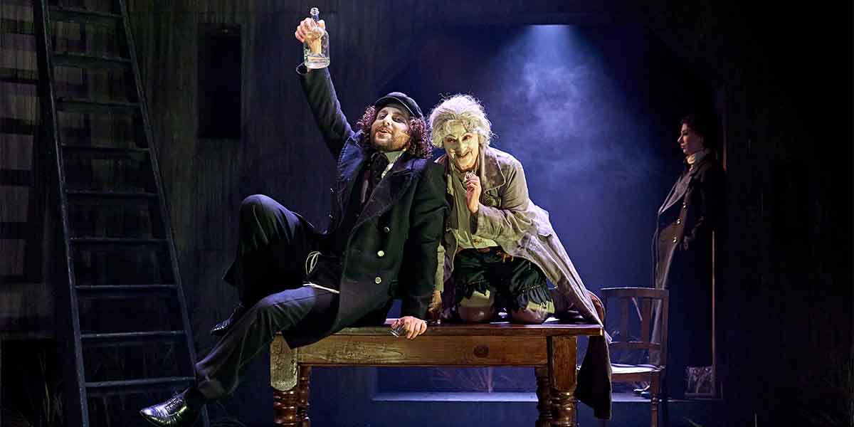 Ashley Margolis as Avigdor raises a bottle, seated on a table beside Evelyn Krape as The Figure.