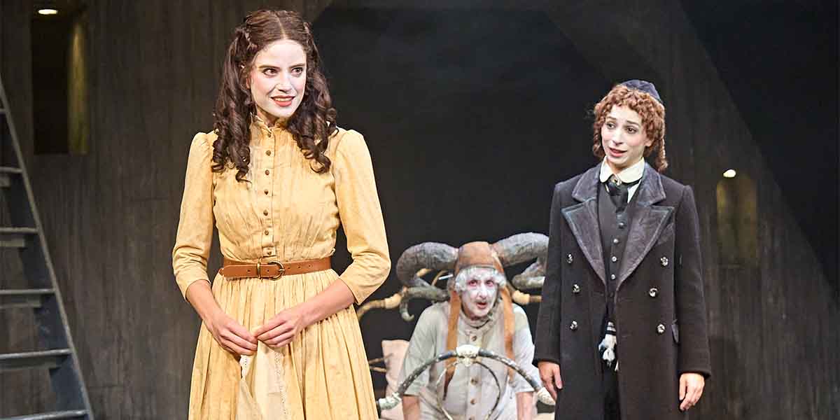 Genevieve Kingsford as Hodes wears a yellow dress, flanked by two characters in a theatrical setting.