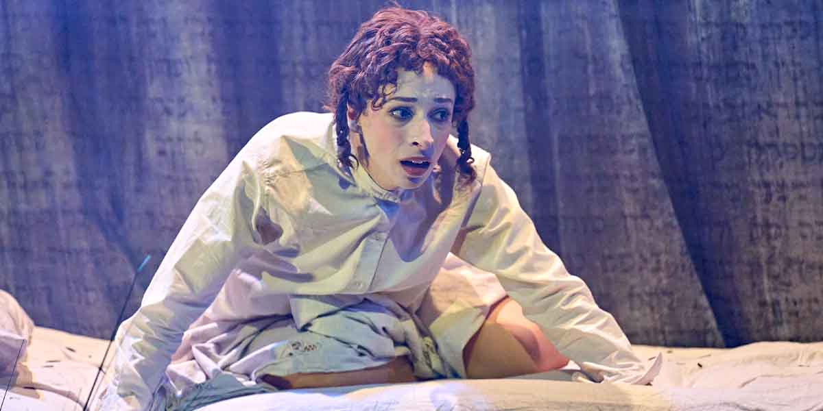 Amy Hack as Yentl, wearing a white shirt, looks surprised while seated on a bed with a textured backdrop.