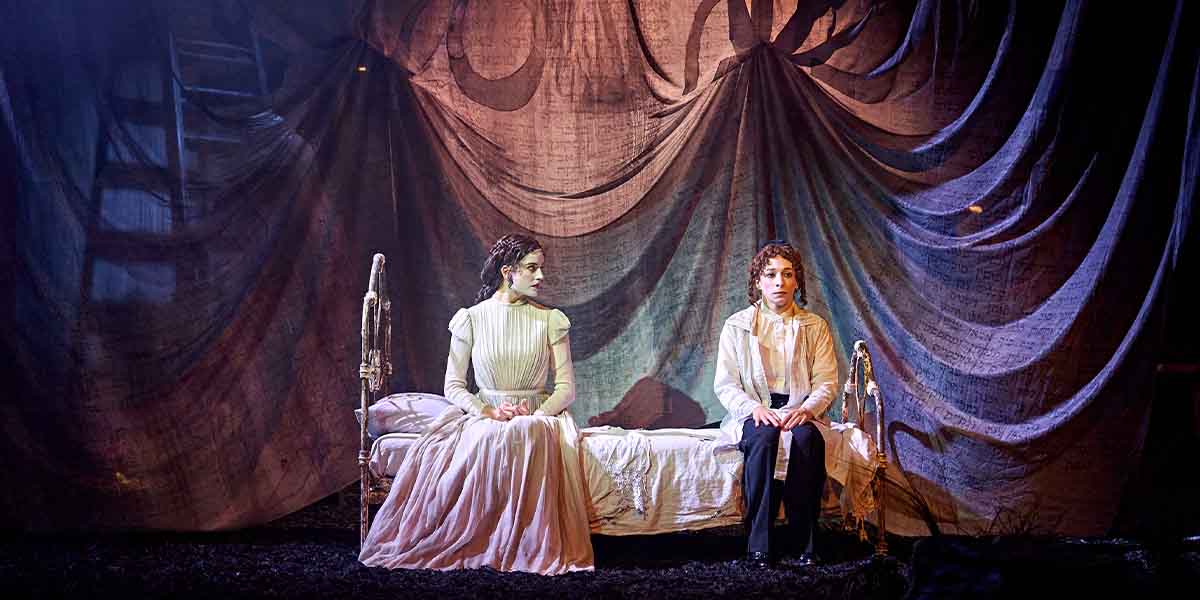 Genevieve Kingsford as Hodes and Amy Hack as Yentl sit on a bed, surrounded by draped fabric.