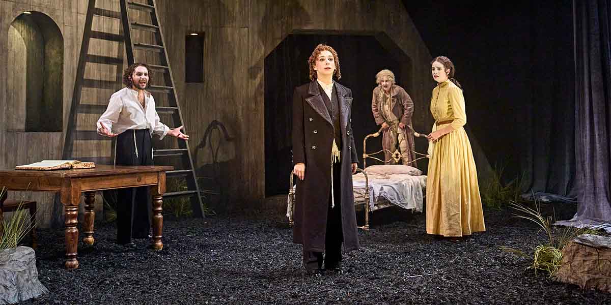 Four actors perform on a dark stage with a wooden table, a ladder, and a bed in the background.