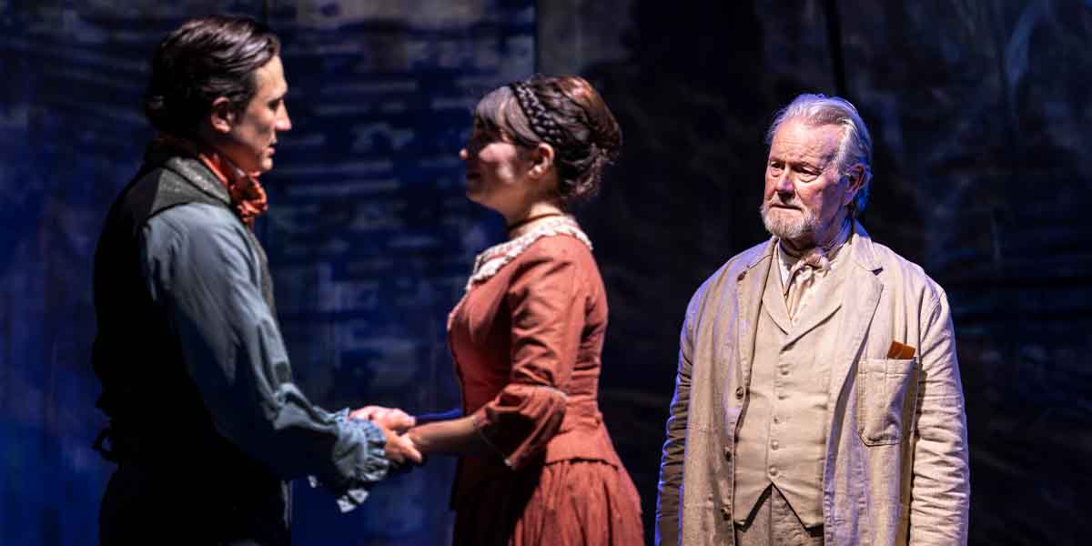 Dean John-Wilson and Brooke Bazarian hold hands, while Jeff Shankley observes, in a theatrical scene.