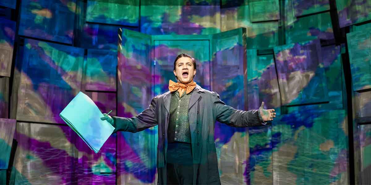 Dean John-Wilson performs energetically on stage, holding a script, against a vibrant, abstract backdrop.