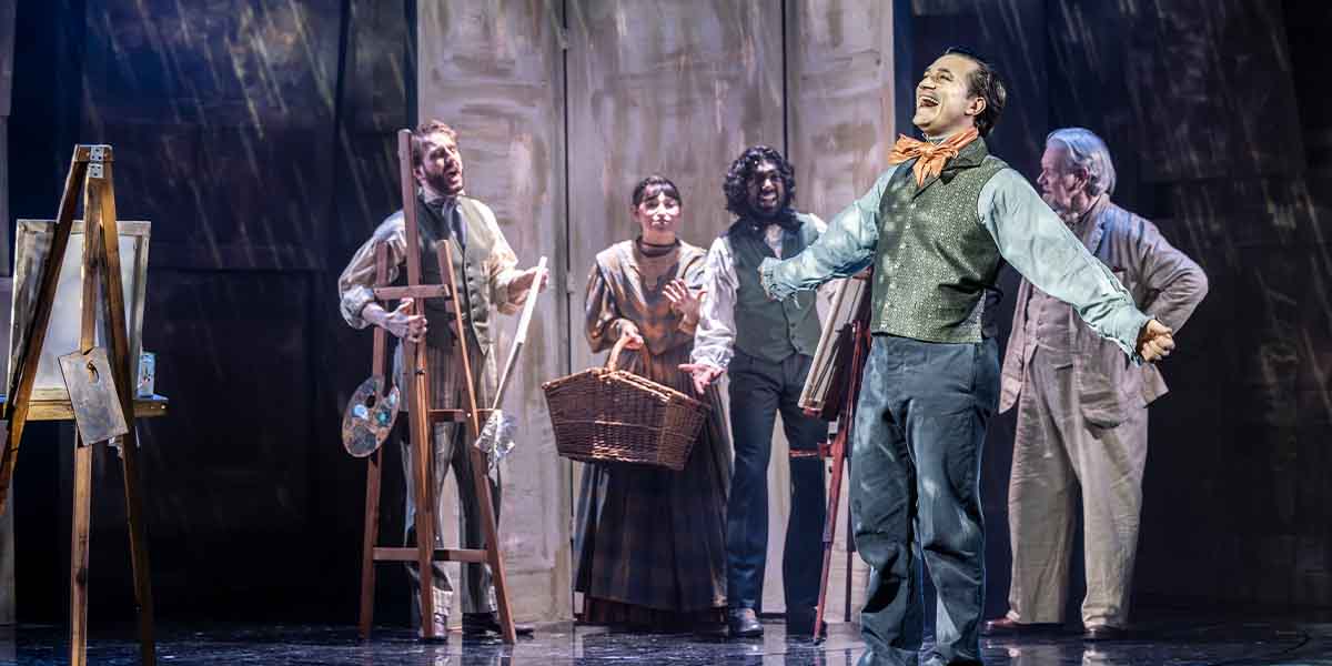 Sam Peggs performs joyfully in front of four actors, with easels and a basket in a theatrical setting.