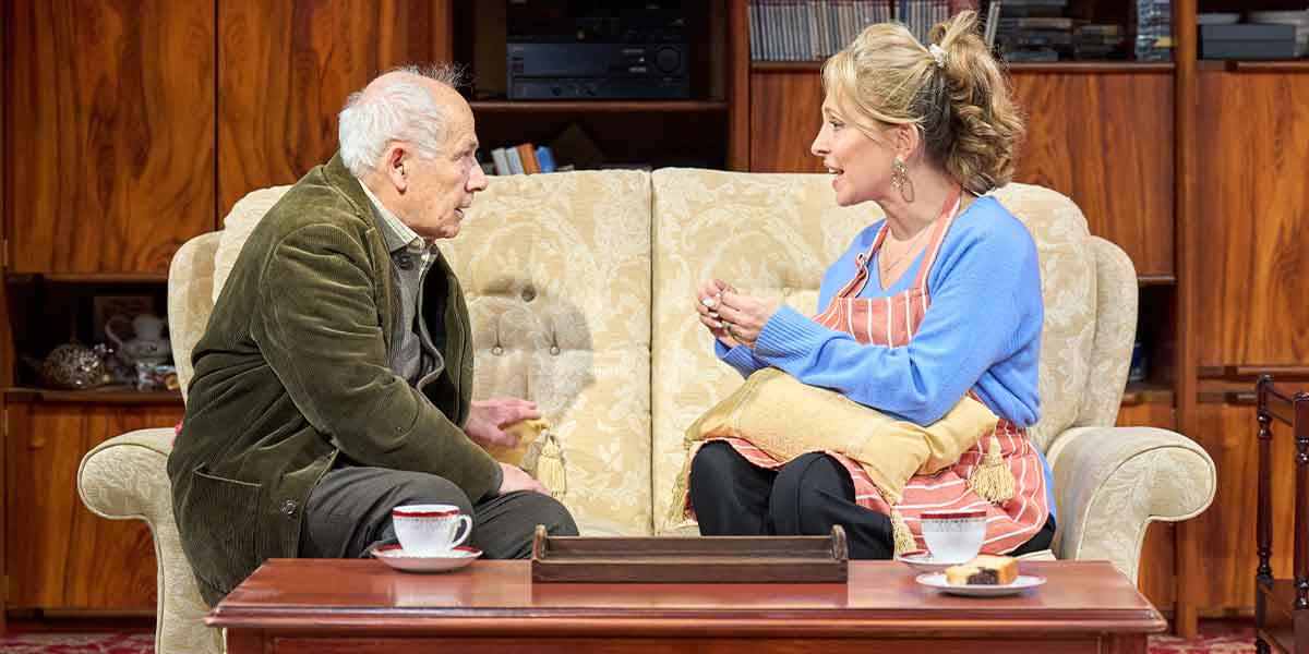 Nicholas Woodeson and Tracy-Ann Oberman sit on a sofa, engaged in conversation, with tea cups nearby.
