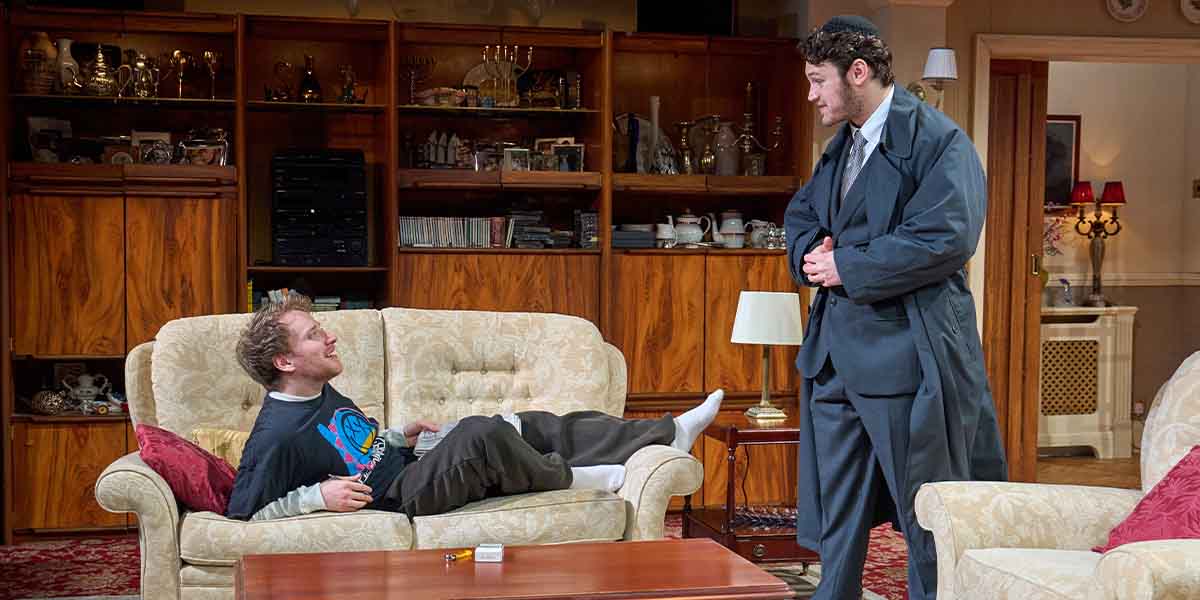 Nitai Levi sits on a sofa, while Alex Zur stands beside him, both engaged in conversation.