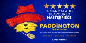Paddington Bear in a red hat and blue coat stands on stage, surrounded by colourful set pieces.