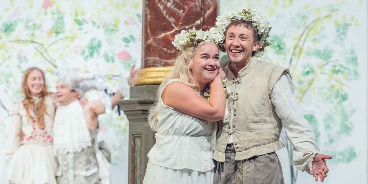 Charlie Josephine as Orlando and Lola Shalam as Rosalind smile, wearing floral crowns, on stage.