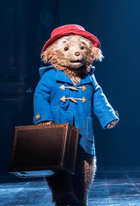 A bear in a blue coat and red hat holds a suitcase, standing on a dimly lit stage.