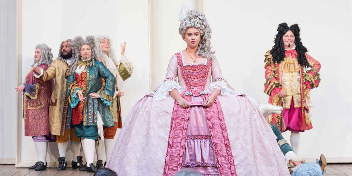 Lola Shalam as Rosalind in a pink gown, surrounded by actors in elaborate historical costumes at Shakespeare's Globe.