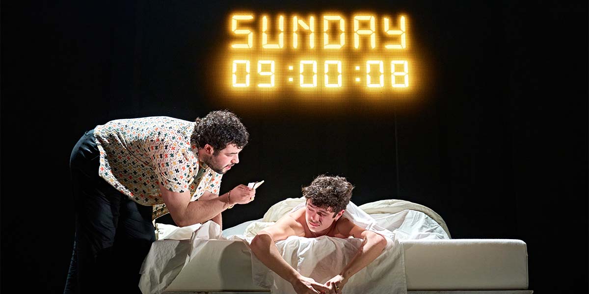 A man in a patterned shirt leans over another man in bed, with a digital clock displaying "SUNDAY 09:00:08.