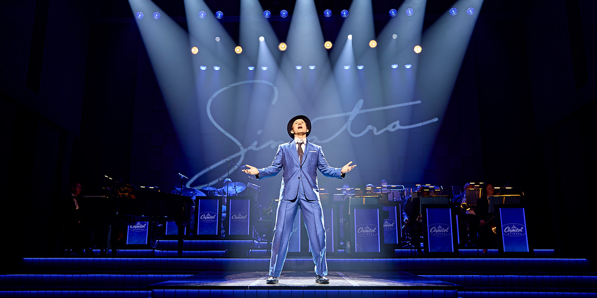Joel Harper-Jackson performs on stage in a suit, surrounded by vibrant stage lighting and a musical backdrop.