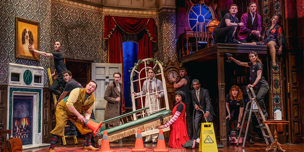 The cast of The Play That Goes Wrong are on stage dressed in various different styles of clothing. All are busy doing an action, some are drilling, some carrying furniture, a window, one is hoovering.