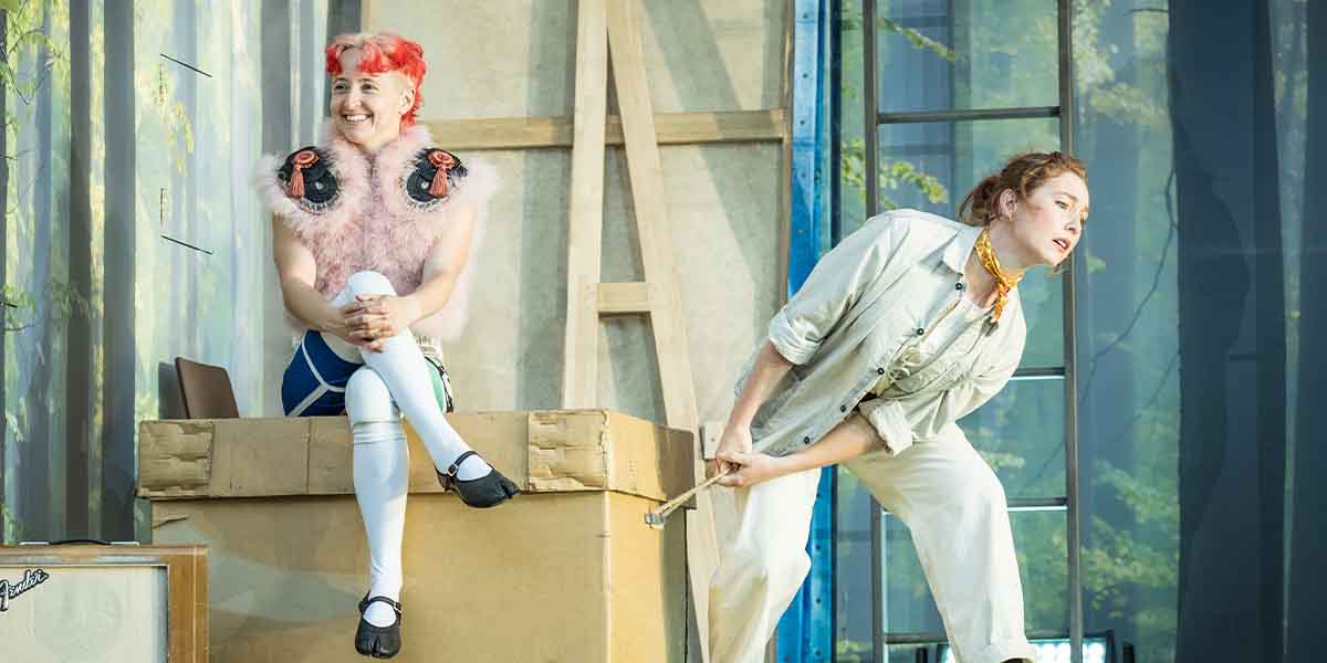 Georgia Bruce as Puck sits on a box, while Harriet Gordon-Anderson as Quince leans forward, holding a stick.