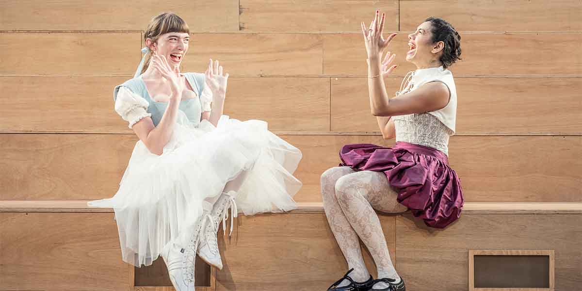 Mary Malone as Helena and Hiftu Quasem as Hermia sit on a wooden bench, laughing joyfully.