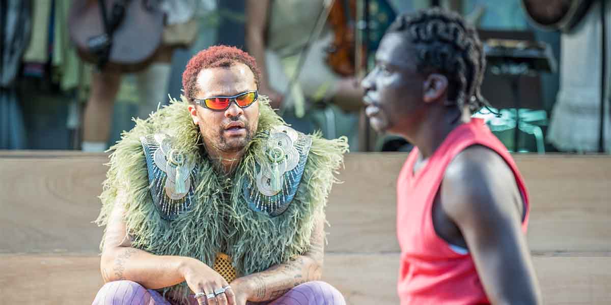 Oliver Huband, wearing a green feathered costume and sunglasses, engages in conversation with Terique Jarrett, in a red shirt.