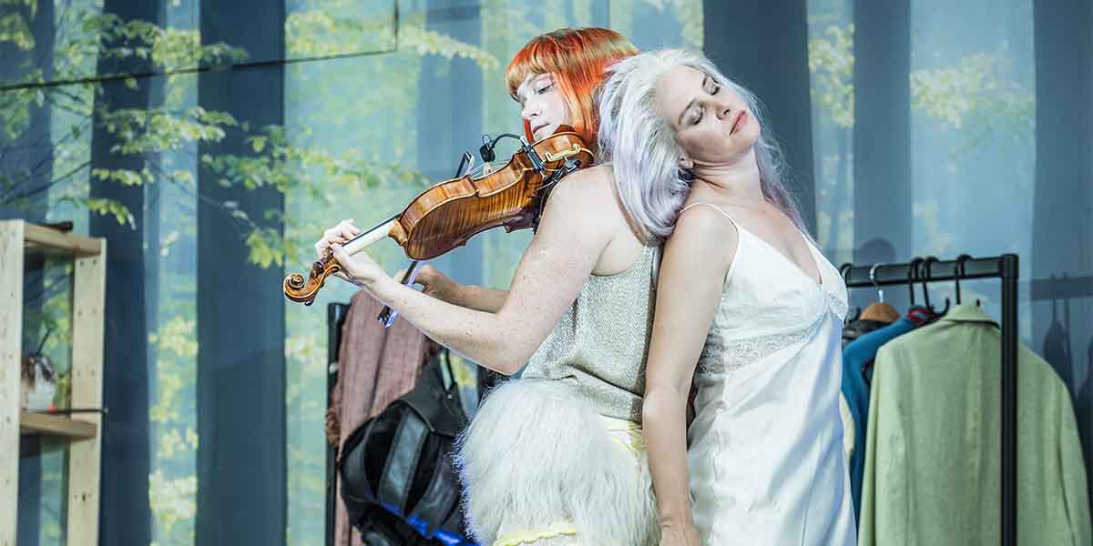 Rori Hawthorn plays the violin while leaning against Jenny Rainsford, who wears a white dress.