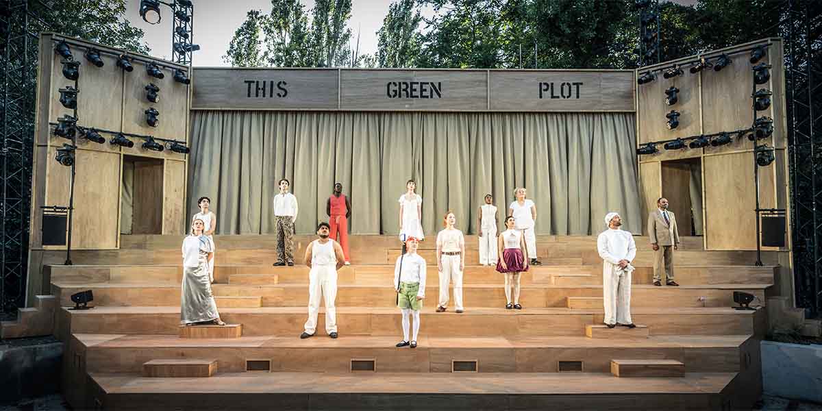 The cast of A Midsummer Night's Dream poses together, wearing vibrant costumes amidst a lush outdoor setting.