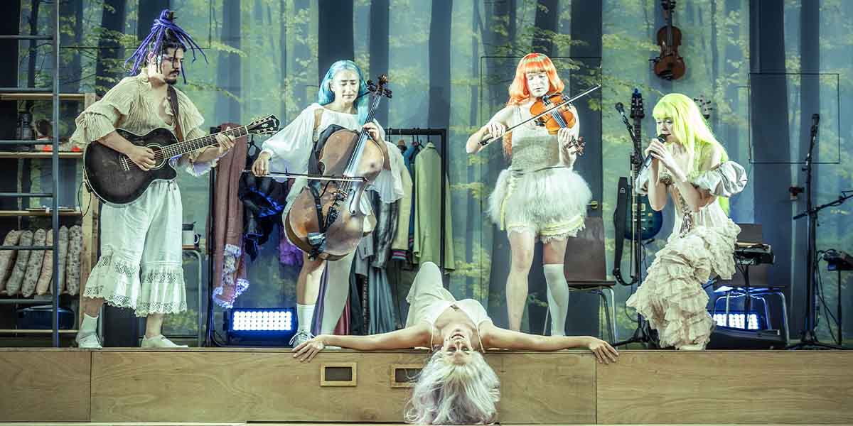 Five performers in vibrant costumes play instruments on stage, with one lying down in the foreground.
