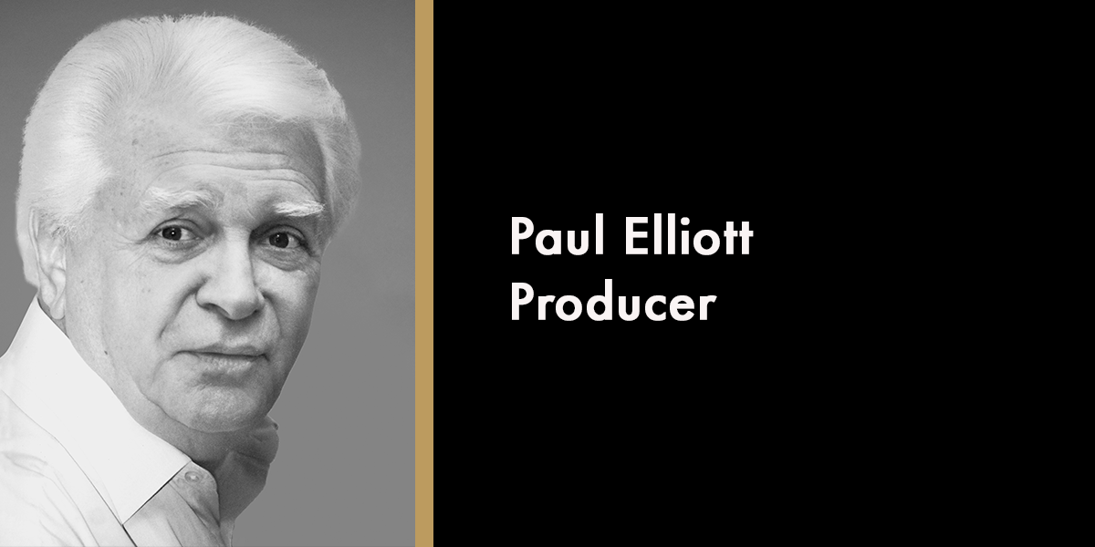 Elliott and Paul stand together, smiling, against a neutral background, both wearing casual clothing.