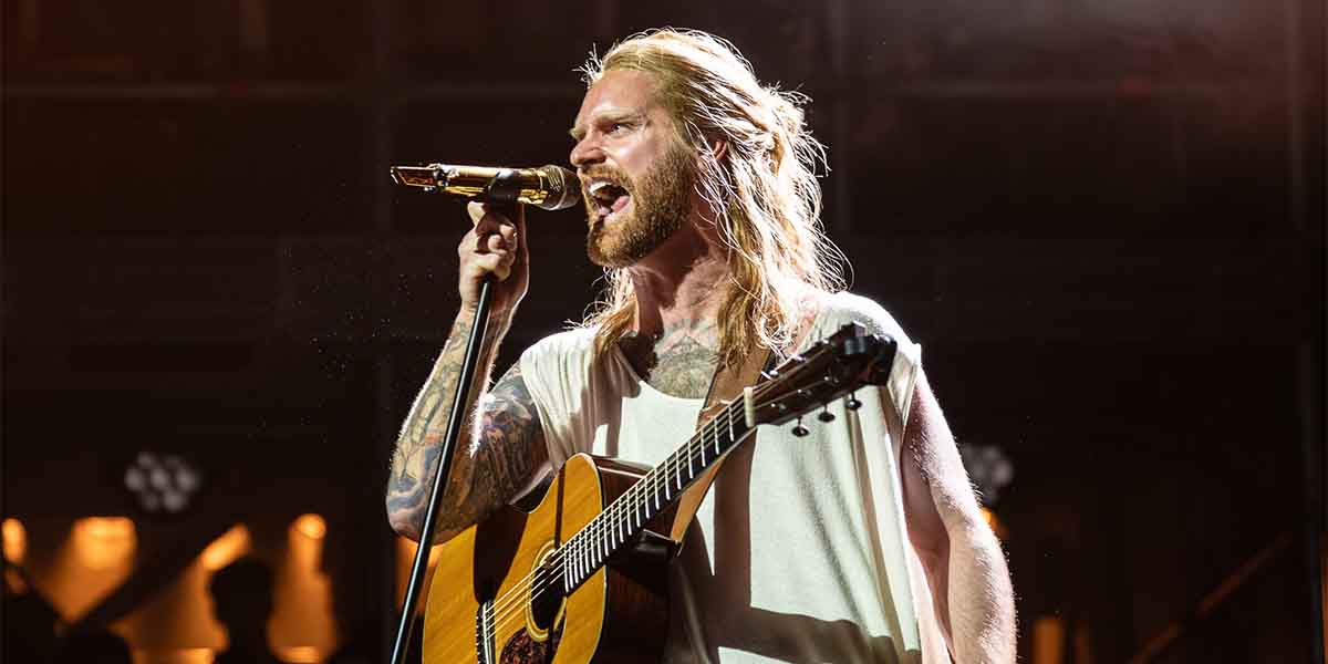 Sam Ryder passionately sings into a microphone while holding an acoustic guitar on stage.