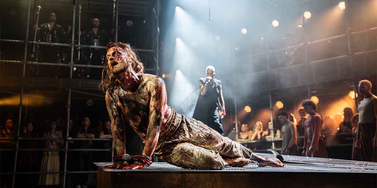 Sam Ryder, covered in dirt and blood, kneels on stage with a microphone, surrounded by performers.