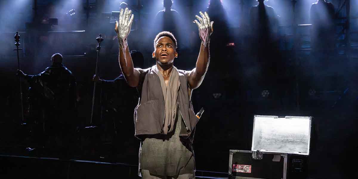 Tyrone Huntley raises his hands, adorned with silver, against a dramatic, illuminated stage backdrop.