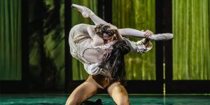 Two performers in ballet attire dance on stage in embrace against a green background.