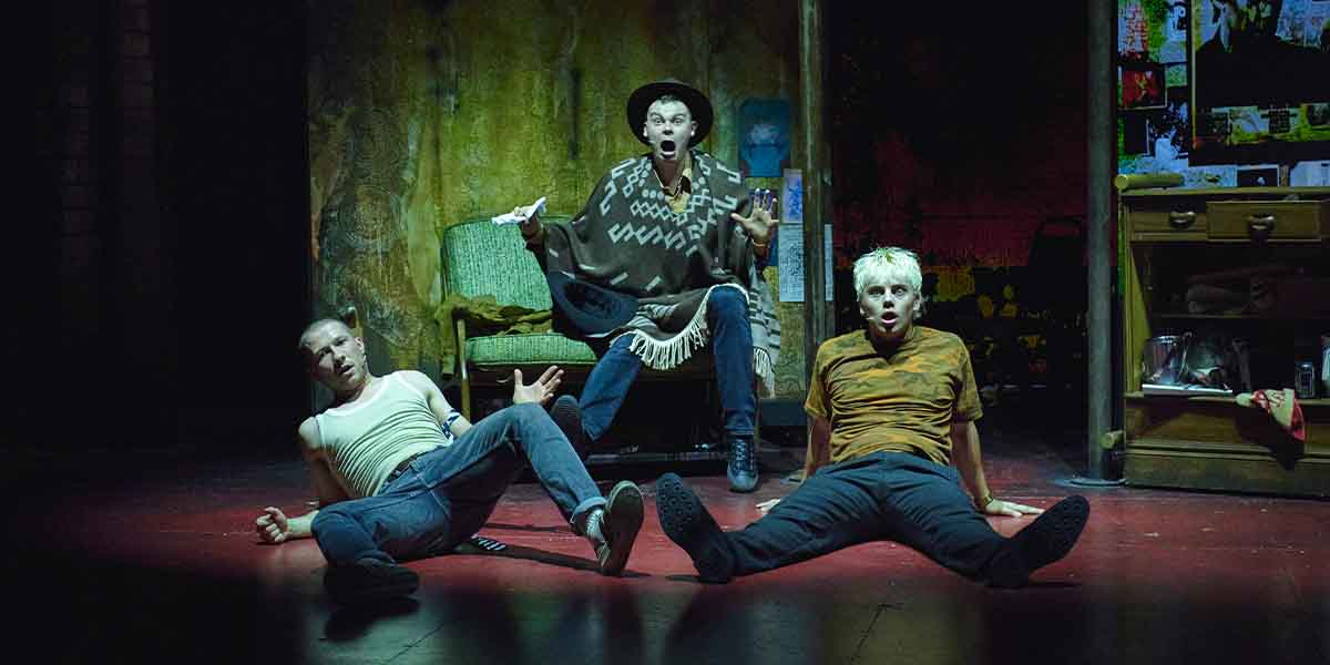 Three actors perform on stage: one in a poncho, two seated, expressing dramatic emotions.
