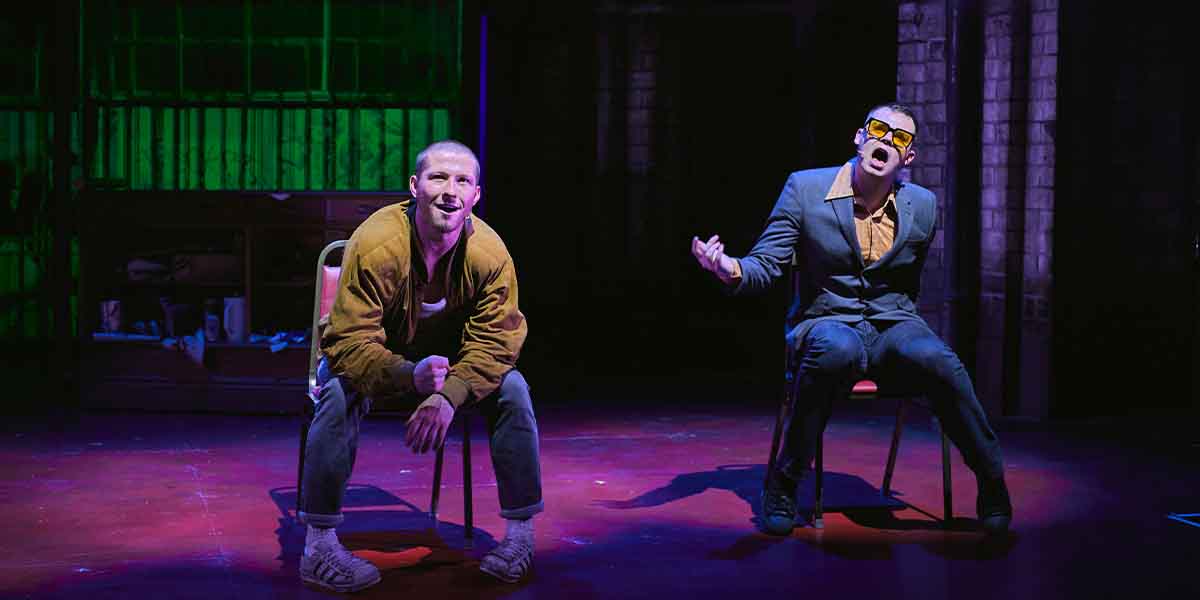 Two male performers sit on chairs, one in a brown jacket and the other in a blue suit, singing.