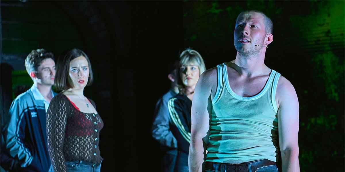 Four performers on stage, with one man in a tank top speaking passionately, dramatic lighting.