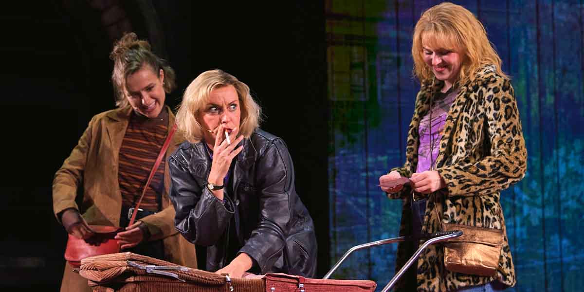 Three women interact on stage; one smokes, while the others examine items in a pram.