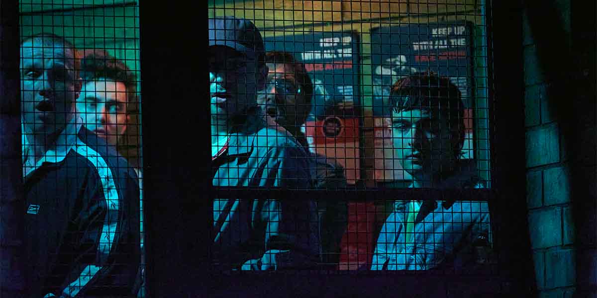 Six men stand behind a wire mesh, illuminated by blue and green lighting, with various expressions.