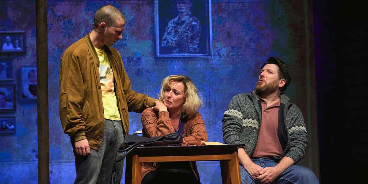 Three actors are seated at a table, engaged in a serious conversation against a textured backdrop.