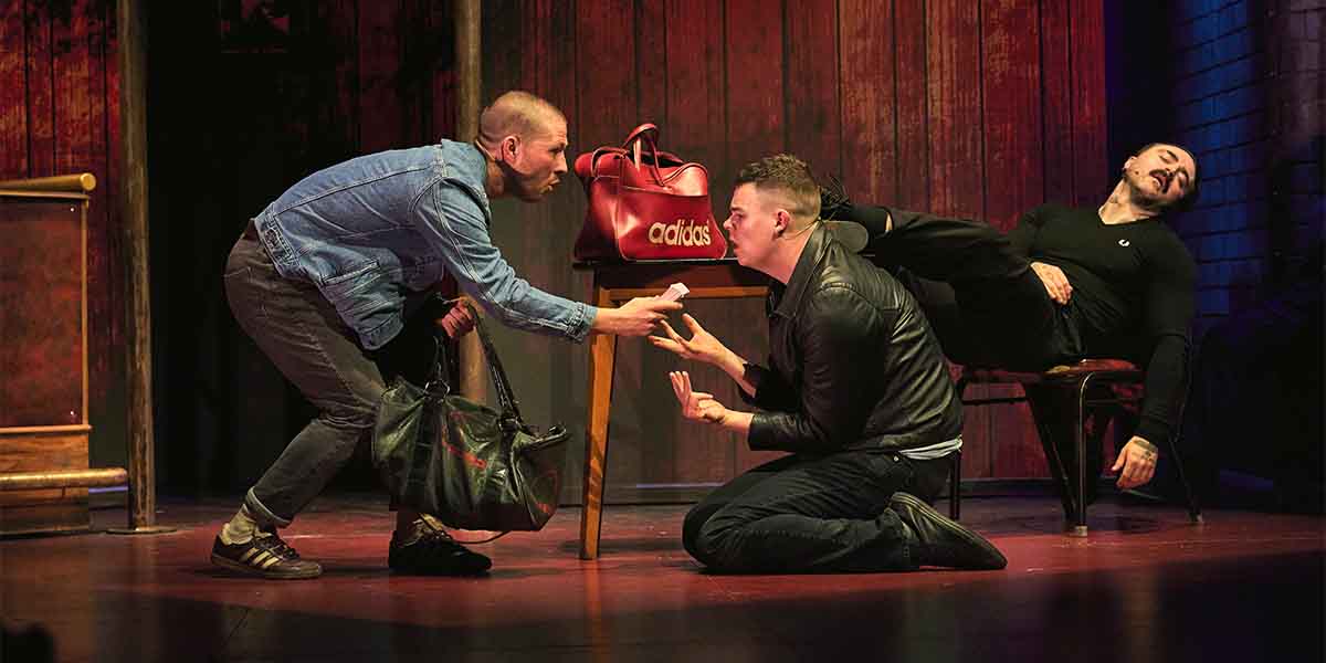Two actors interact on stage, one kneeling with a bag, while another leans on a chair.