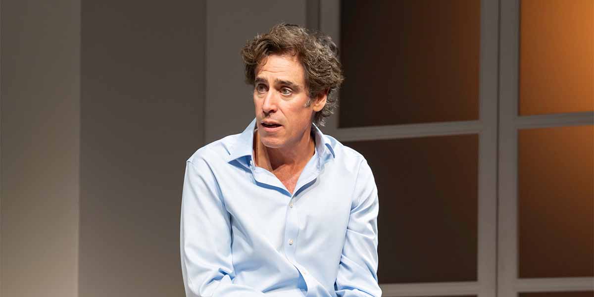 A person with curly hair, wearing a light blue shirt, appears thoughtful while seated on stage.