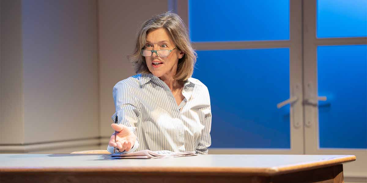 A person with glasses gestures while speaking at a table, wearing a striped shirt against a blue background.