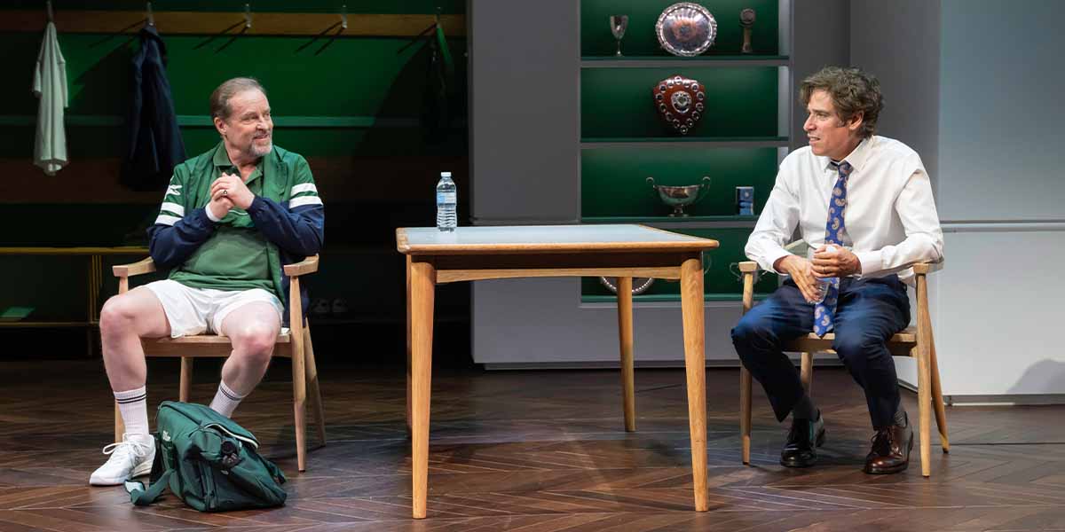 Two people sit at a table; one in sportswear, the other in a shirt and tie, both engaged in conversation.