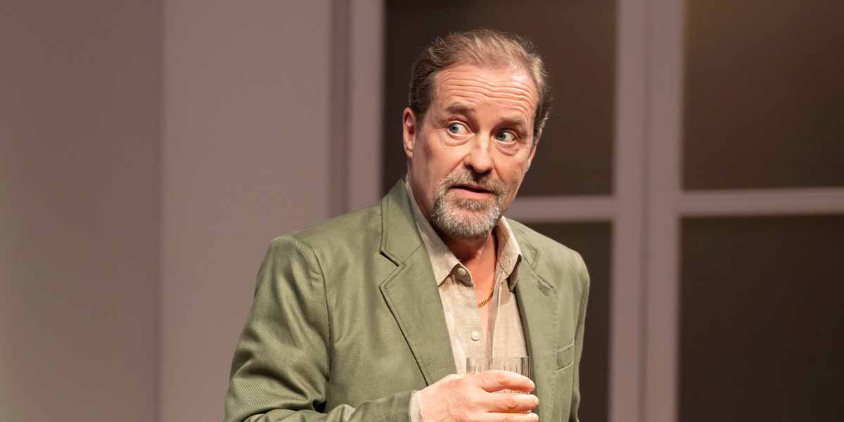 A person with a beard and surprised expression holds a glass, wearing a light green jacket.