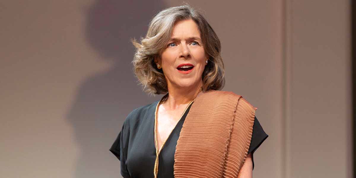 A person with wavy hair looks surprised, wearing a black top and a textured brown shawl.
