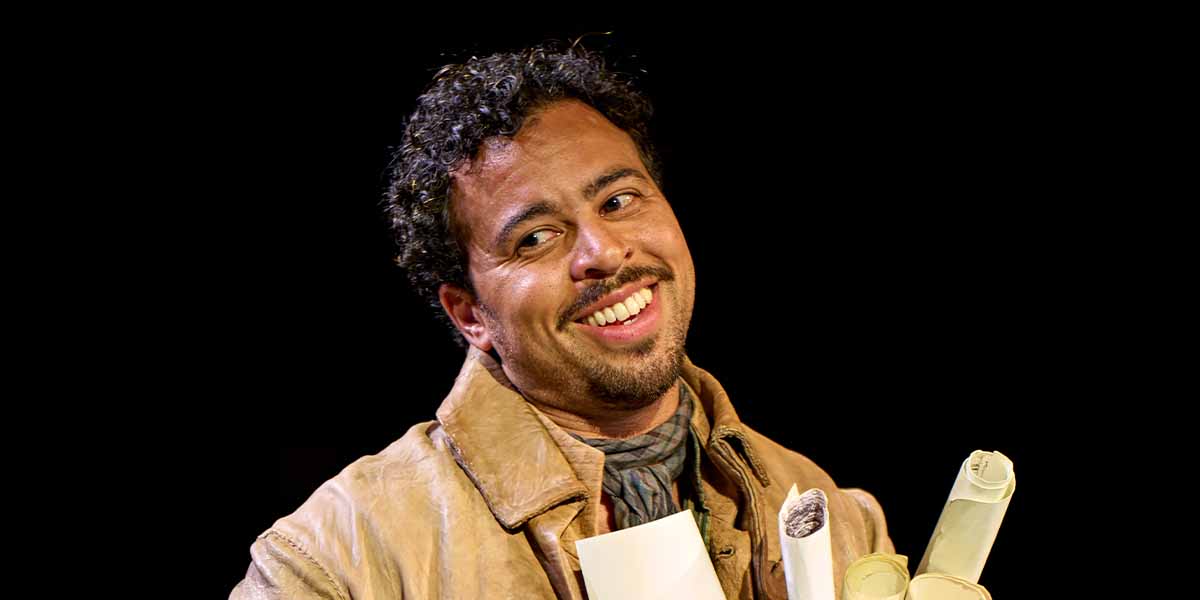 Aaron Anthony as Richard Noakes smiles while holding scrolls, wearing a beige coat against a dark background.