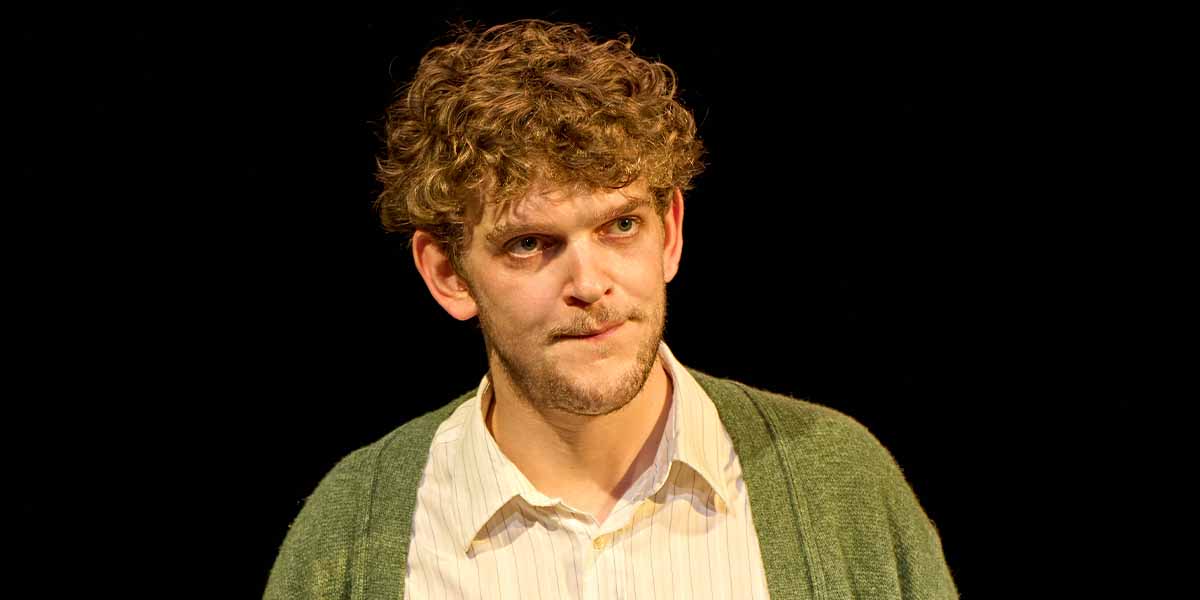 Angus Cooper as Valentine Coverly, wearing a white shirt and green cardigan, with a serious expression.