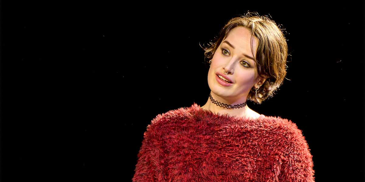 Holly Godliman as Chloe Coverly, wearing a red fluffy sweater, with a black background.
